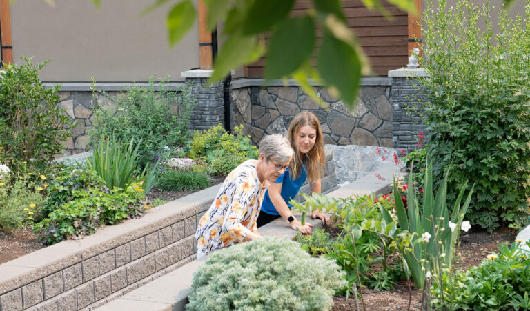 A female senior resident at Origin at Spring Creek and a Life Enrichment Coordinator in a blue uniform look at flowers and greenery in a garden space at Spring Creek